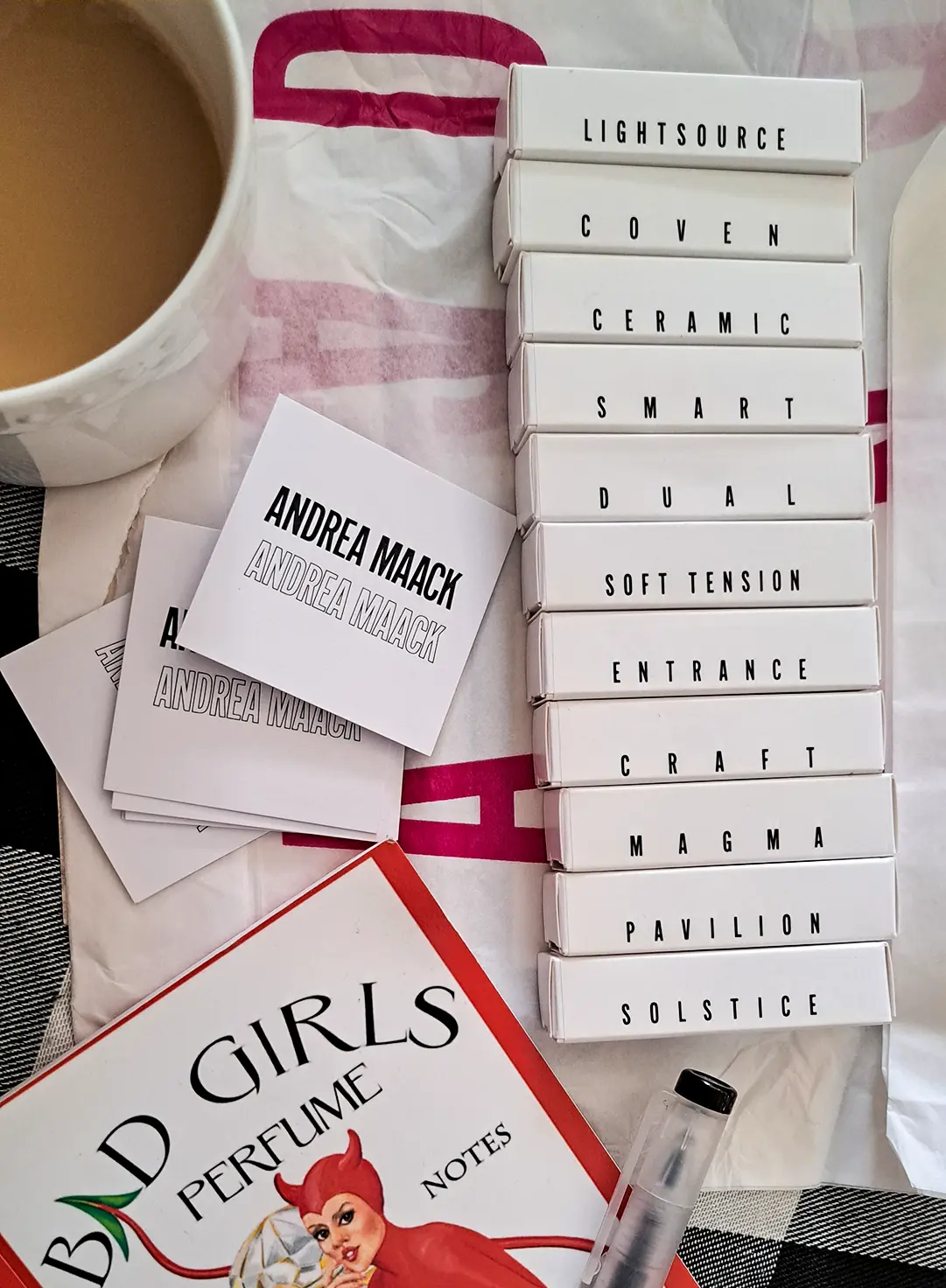 Andrea Maack discovery set featuring sample boxes for Lightsource, Coven, Ceramic, and more, alongside the 'Bad Girls Perfume Notes' book and a cup of coffee.