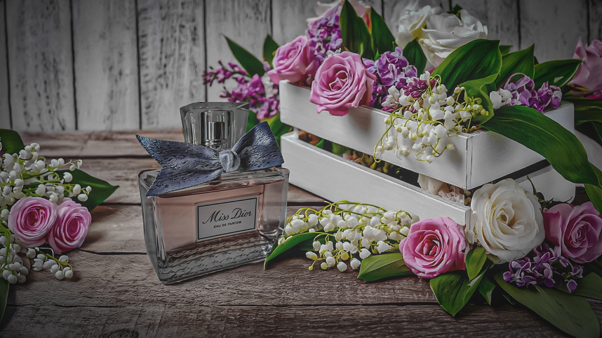 A bottle of Miss Dior Eau de Parfum adorned with a blue ribbon, nestled amongst a variety of pink and white roses, lily of the valley, and purple lilac in a rustic wooden setting.