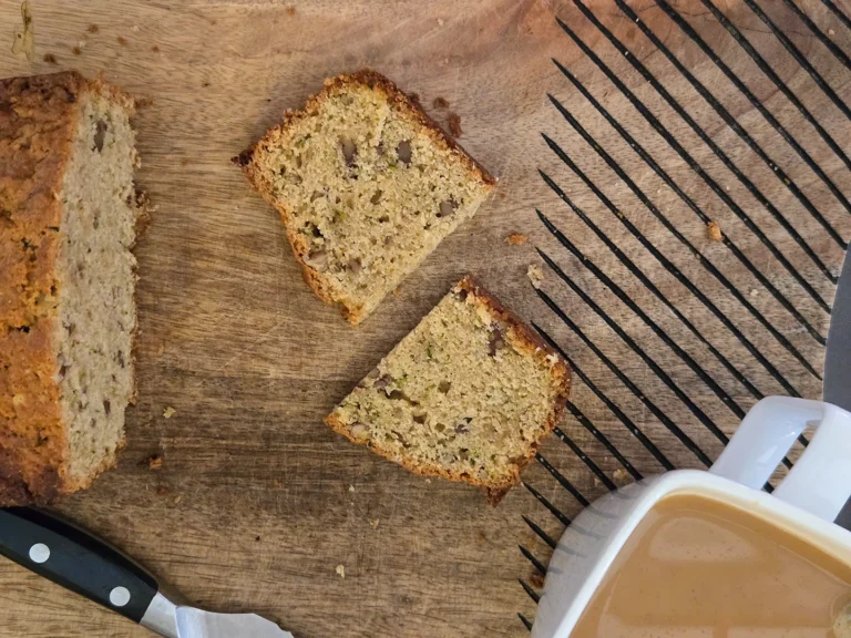An overhead view of a partially sliced loaf of zucchini pecan bread on a wooden cutting board. Two slices are laid out next to a bread knife and a cup of coffee.