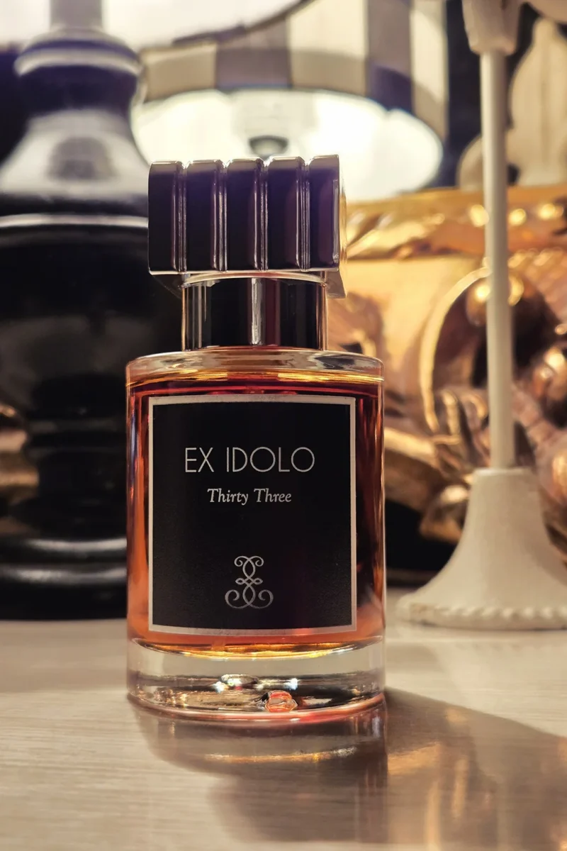 A bottle of Ex Idolo Thirty-Three perfume sits on a wooden surface, with warm, ambient lighting from lamps in the background creating a luxurious and intimate mood.