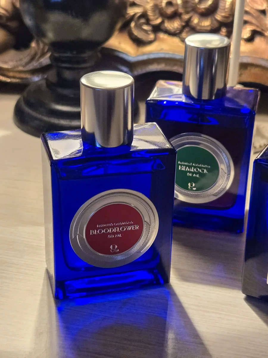 A close-up of the cobalt blue Parfums Quartana Bloodflower bottle, illuminated by cool, dramatic light, with the Hemlock bottle visible in the background.