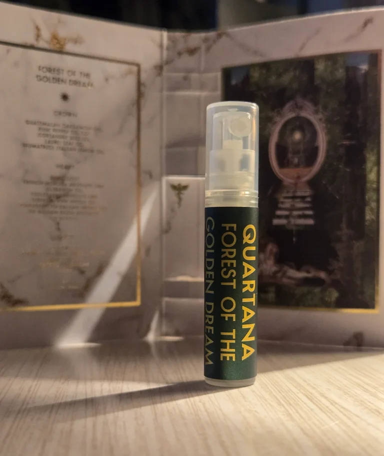 A close-up of the Quartana Forest of the Golden Dream sample spray vial, standing upright in front of its open, blurred-out sample card.