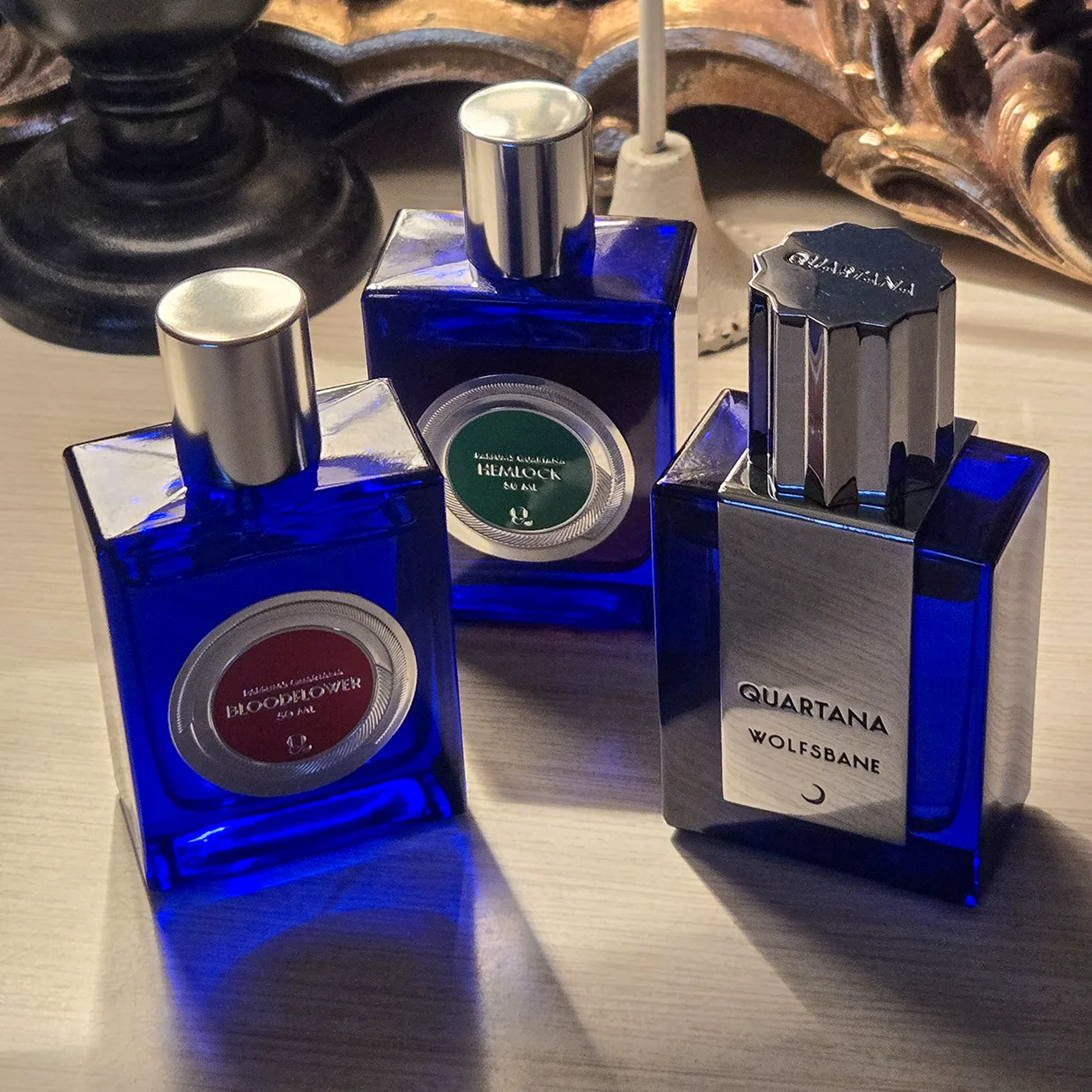 Three cobalt blue bottles from the Parfums Quartana Les Potions Fatales collection, featuring Bloodflower, Hemlock, and Wolfsbane, arranged artfully with dramatic shadows.