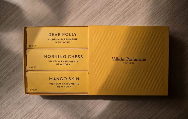 A yellow Vilhelm Parfumerie discovery set box, open to show 2ml samples of Dear Polly, Morning Chess, and Mango Skin, resting on a wooden surface in warm, dramatic lighting.