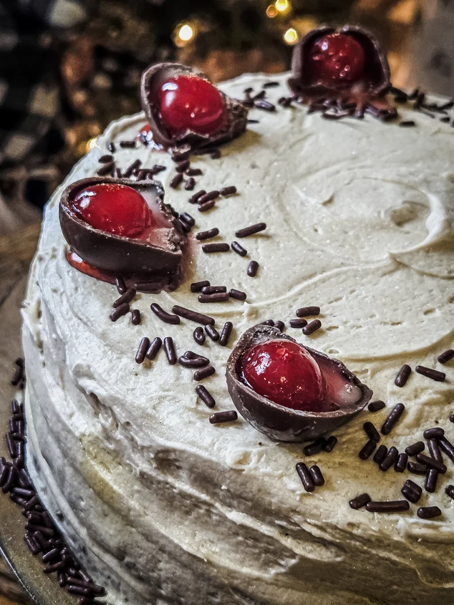 Extreme close-up of the top of a frosted cake featuring dark chocolate sprinkles and halved maraschino cherry cordials with liquid centers.
