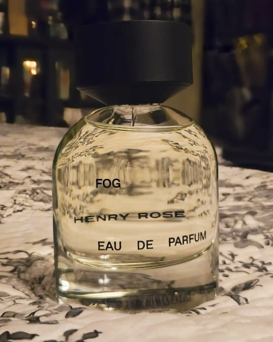 A clear glass bottle of Henry Rose "Fog" Eau de Parfum with a matte black cap, centered on a floral patterned surface.